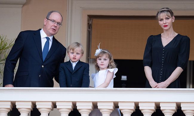 albert and charlene dresssed in formal navy attire on either side of a young boy and girl on a balcony stone balustrades