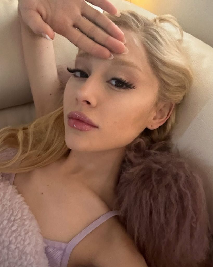 Ariana Grande reclines in true Glinda fashion wearing a soft pastel slip. Her hair is blonde and her arm is raised above her head. She has full pink lips, French manicured nails and long lashes - typical Glinda makeup