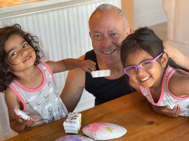 two little girls wearing matching pink and grey vest tops lean together on either side of their adopted father who also wears a vest and they are laughing as they look at the camera