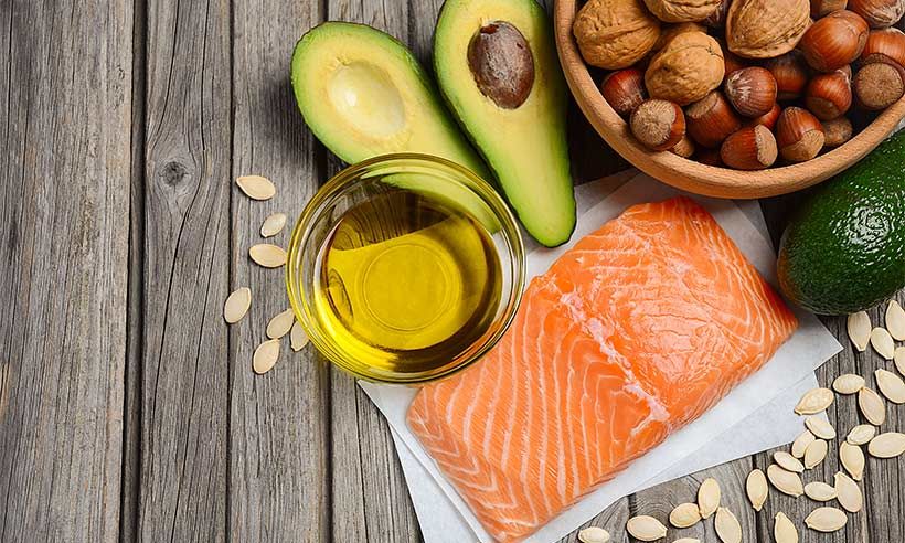 a fillet of raw salmon a glass of oil an open avocado and a bowl of nuts with sunflower seeds are placed on a wooden table as an example of omega rich food