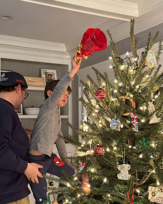 brian lifts his young son up in order to place a red angel at the top of a christmas tree which is decorated with lights and toy ornaments