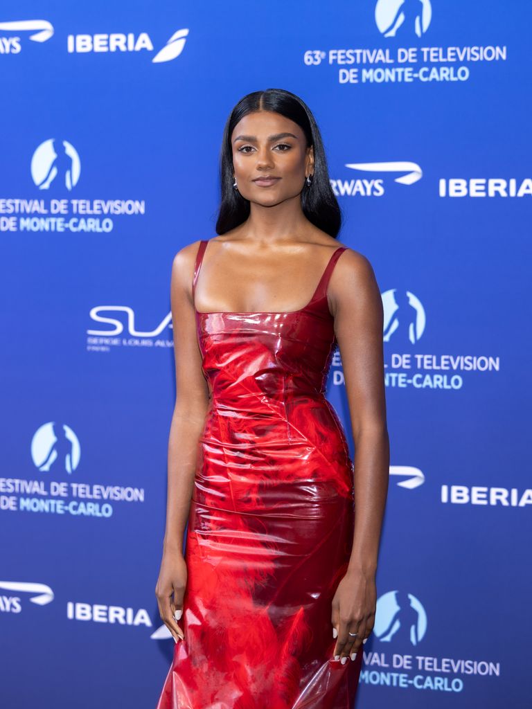 Simone Ashley stands on a navy blue carpet against a backdrop reading "63rd Festival de Télévision de Monte-Carlo." She is wearing a fitted, glossy red dress with thin shoulder straps and a unique balloon-style skirt that cinches in at the hem. The fabric has a highly reflective, lacquered texture that gives it a modern, high-shine appearance. Her long black hair is sleek and straight, parted in the middle. She pairs the dress with open-toe heels with transparent straps and clear block heels, giving her an almost floating appearance.