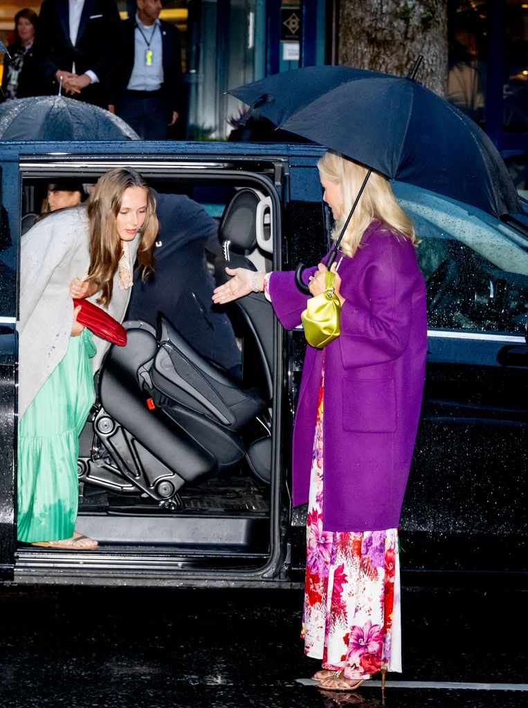 Crown Princess Mette-Marit in a purple coat holding an umbrella and extending an arm to Princess Ingrid Alexandra