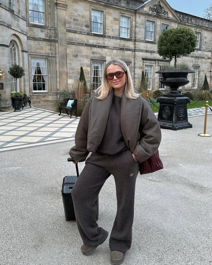 Molly-Mae stands in front of a grand stone building, wearing a brown bomber jacket and cozy loungewear, pulling a suitcase with a burgundy woven tote on her shoulder.