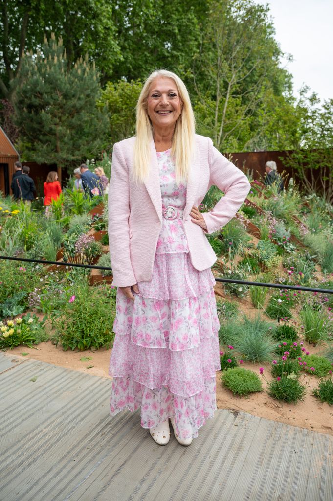 Vanessa Feltz attends Chelsea Flower Show 2025 in a layered pink floral tiered dress, pale pink boucle blazer and white studded clogs, posing in front of a garden landscape.