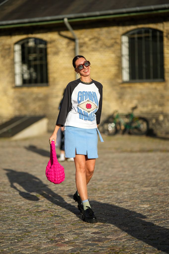 A guest wears brown sunglasses, gold earrings, a gold large necklace, a black and white bicolored with blue and red print pattern long sleeves t-shirt from Ganni, a neon pink quilted pattern handbag, a blue large fringed knees short skirt, blue / green / beige tulle embroidered pattern socks, black shiny leather derbies shoes , outside Ganni, during the Copenhagen Fashion Week Spring/Summer 2024