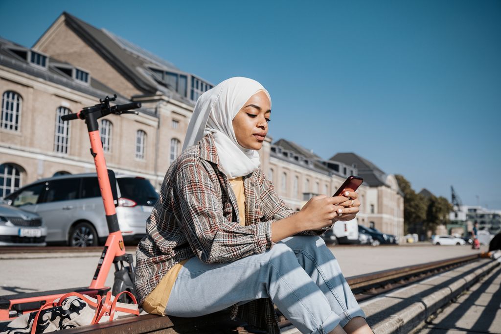 young muslim woman with hijab sitting at riverside in berlin and tapping at  mobile