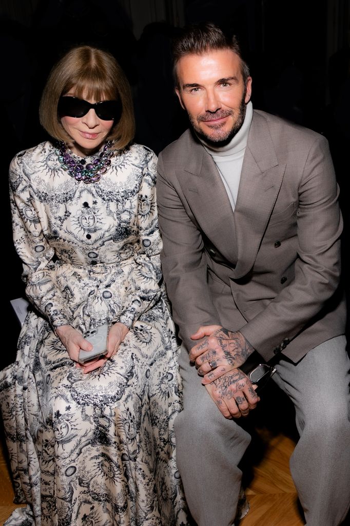 Anna Wintour and David Beckham sit on the front row of a Victoria Beckham fashion show. David is wearing a grey blazer, trousers and polo knit. 