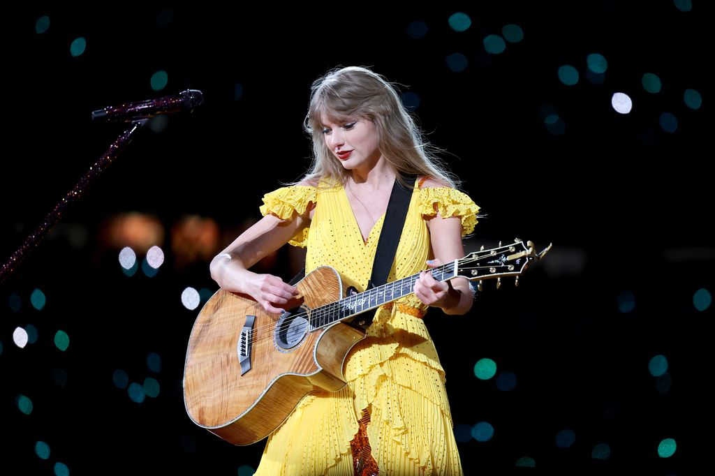 Taylor Swift plays guitar as she wears yellow jumpsuit