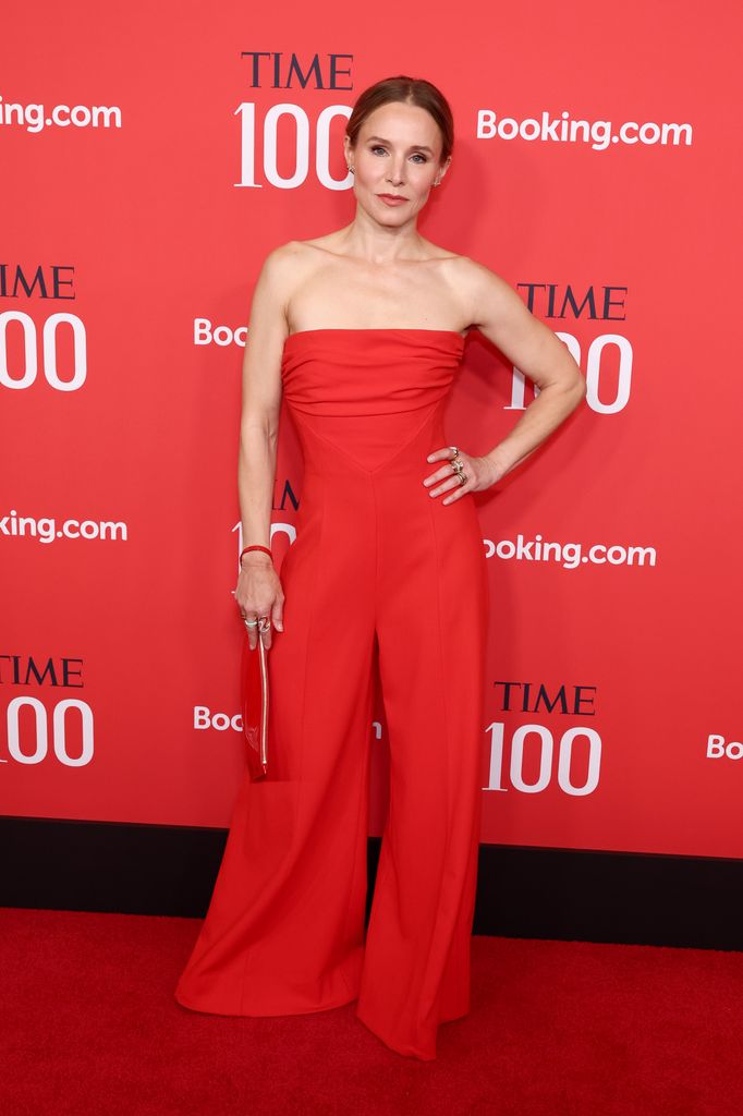 Kristen Bell wears a red strapless jumpsuit and poses on the red carpet with one hand on her hip