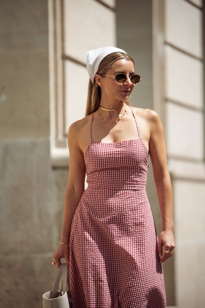 Julia Comil porte une robe maxi à carreaux rouges et blanc avec des sangles, une écharpe à tête blanche, des lunettes de soleil et un sac beige et blanc pendant la Haute Couture Fall / Winter 2025/2026 dans le cadre de la Fashion Week de Paris 
