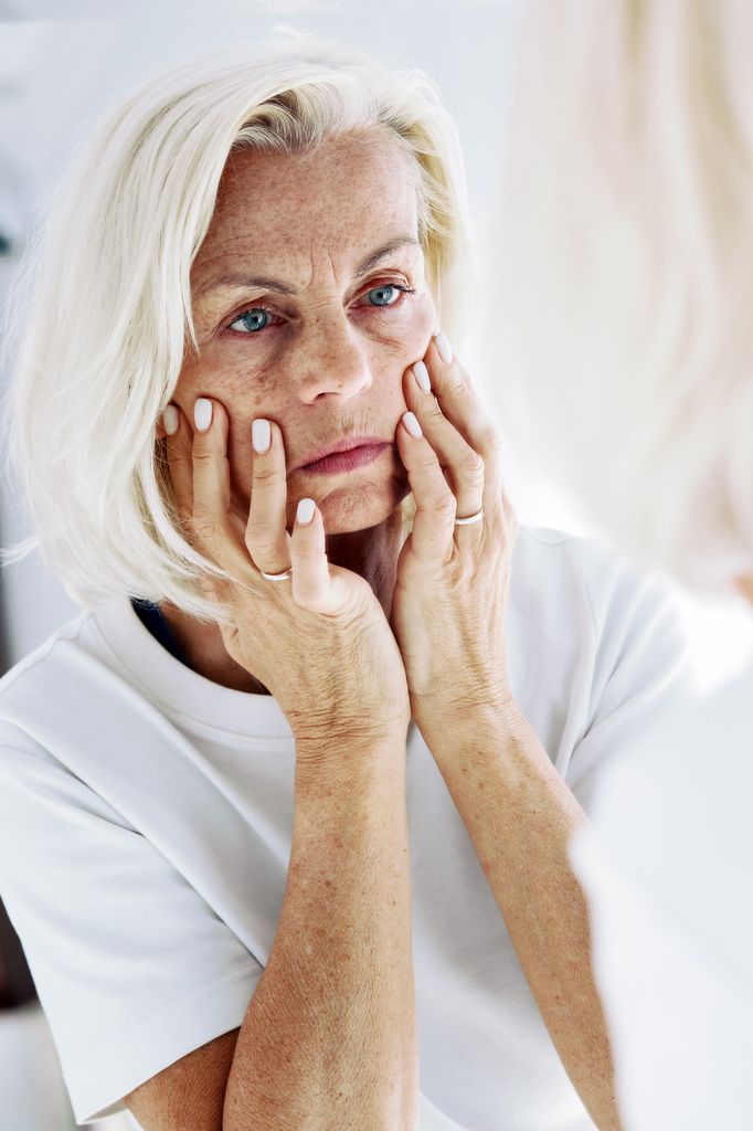 Senior woman with blonde hair examining, touching her facial wrinkles with concerned expression.