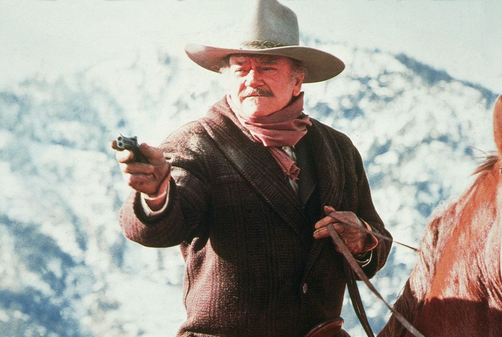 American actor John Wayne (1907 - 1979) aims a gun while riding horseback in a scene from 'The Shootist'