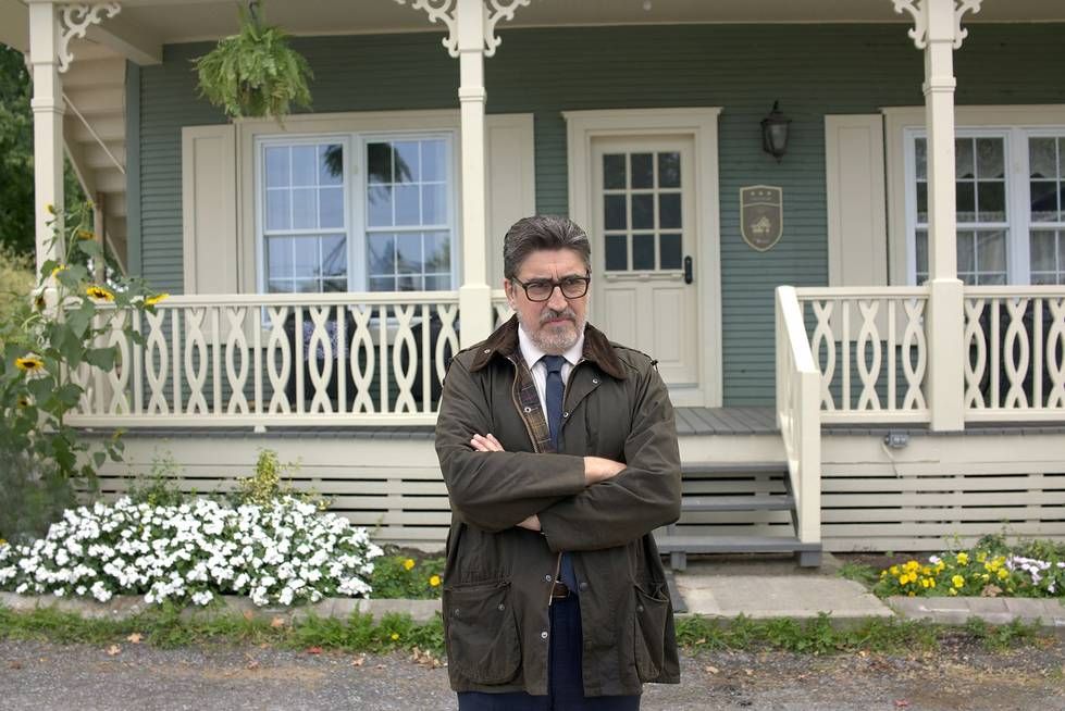 Alfred Molina with his arms crossed standing in front of a house