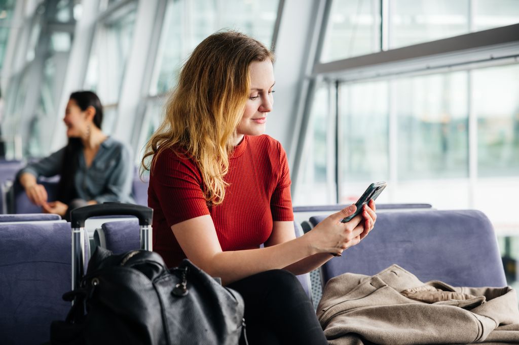 A woman wearing a red shirt and with long, strawberry hair using her smartphone to pass the time while waiting in an airport lounge for a flight.