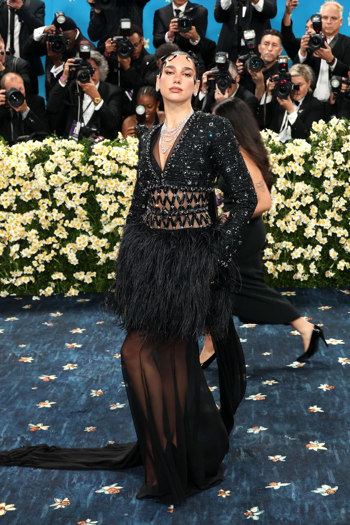 Dua Lipa wears a black cropped jacket paired with a black dress with cut out detail on the waistline, feathers on the hem and a sheer mesh skirt.