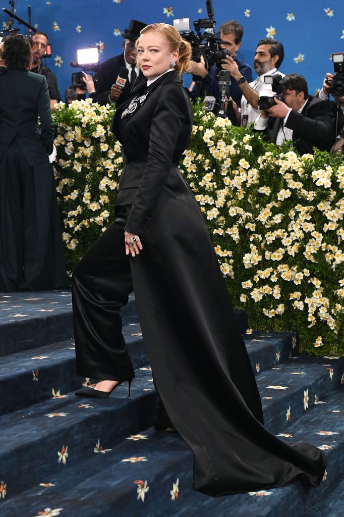 Sarah Snook wears a long black maxi jacket teamed with satin trousers and black heels.