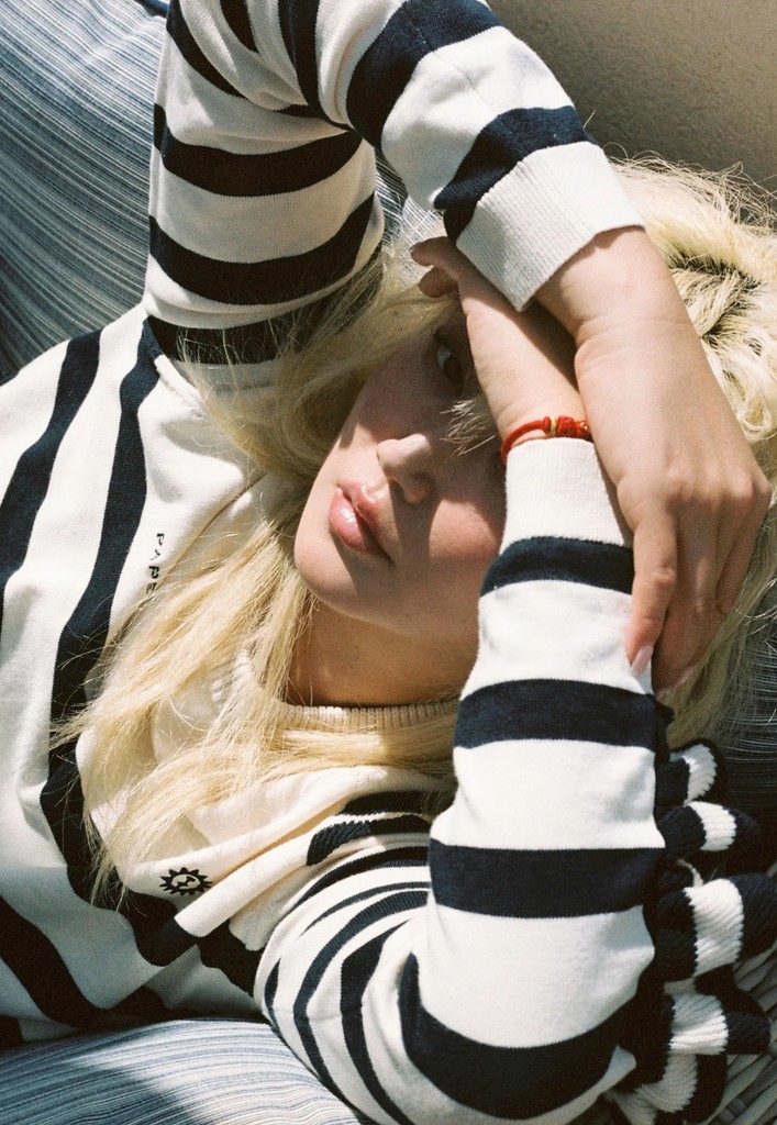 Betty Bachz lounges in a cream and navy striped Paper London x AREV jumper, resting her arm across her forehead. The shot is sun-dappled and moody, exuding Riviera cool.