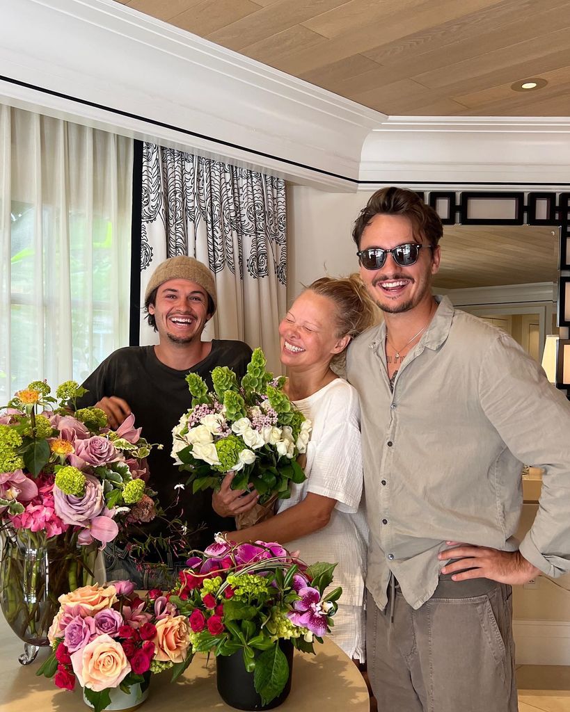 Pamela Anderson riant à la maison en tenant des fleurs à côté de deux fils