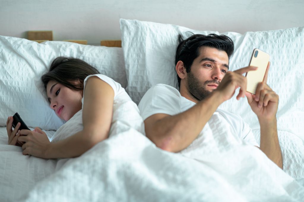 Shot of a young couple using their cellphones in bed at evening back to back .
