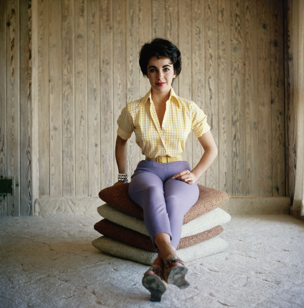 Undated photograph of actress Elizabeth Taylor, sitting on a stack of pillows, wearing purple pants and a yellow shirt, and smiling.