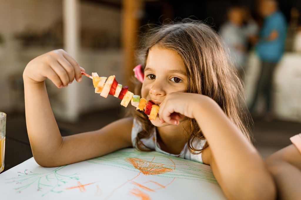 Small girl eating fresh fruit kebab.