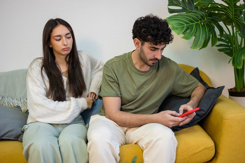 Young couple sitting on sofa in apartment living room, girlfriend looks angry while her boyfriend is texting a girl.