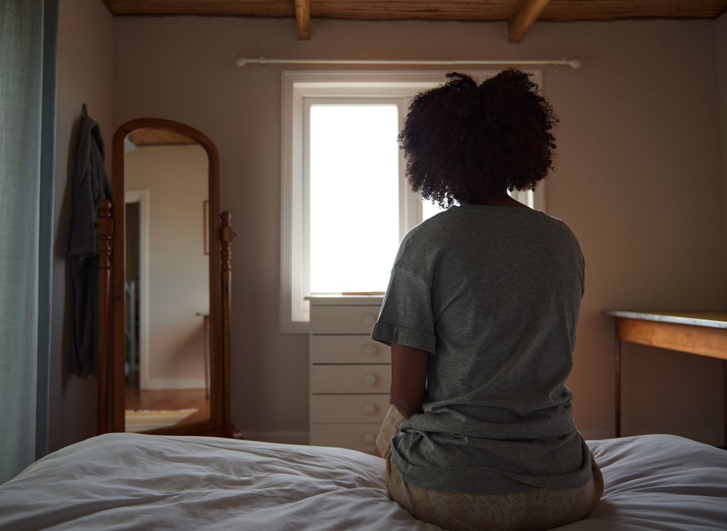 Rear view of a young woman  sitting on her bed and looking out through a window