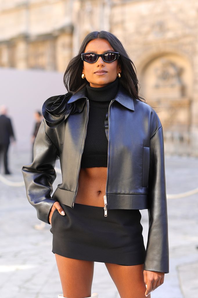 Simone Ashley wears a cropped leather jacket, black top and black mini skirt. She poses with one hand in her pocket and wears sunglasses and a small pair of gold hoop earrings.