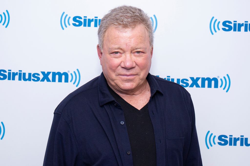 William Shatner in a navy shirt standing in front of SiriusXM logo sign