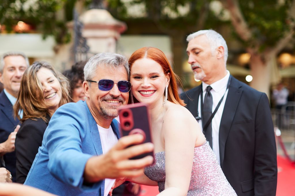A man and woman take a selfie with other people around them at the red carpet event.