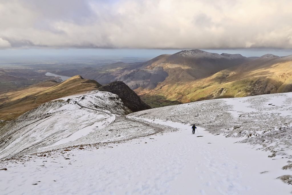 A solitary hillwalker is visible on the snow covered route down the Llanberis Path from the summit of Snowdon, Snowdonia National Park. Llanberis is visible in the distance.