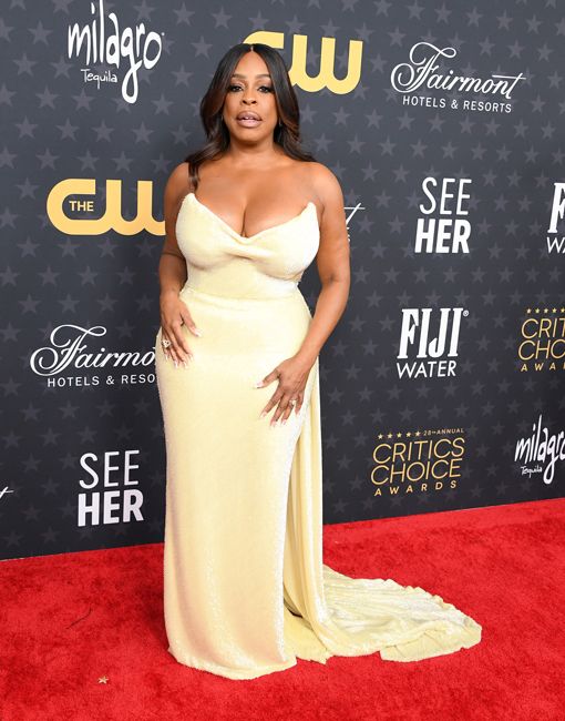 niecy stands on a red carpet in her strapless form fitting sequin gown which has a train and appears light yellow in the daylight