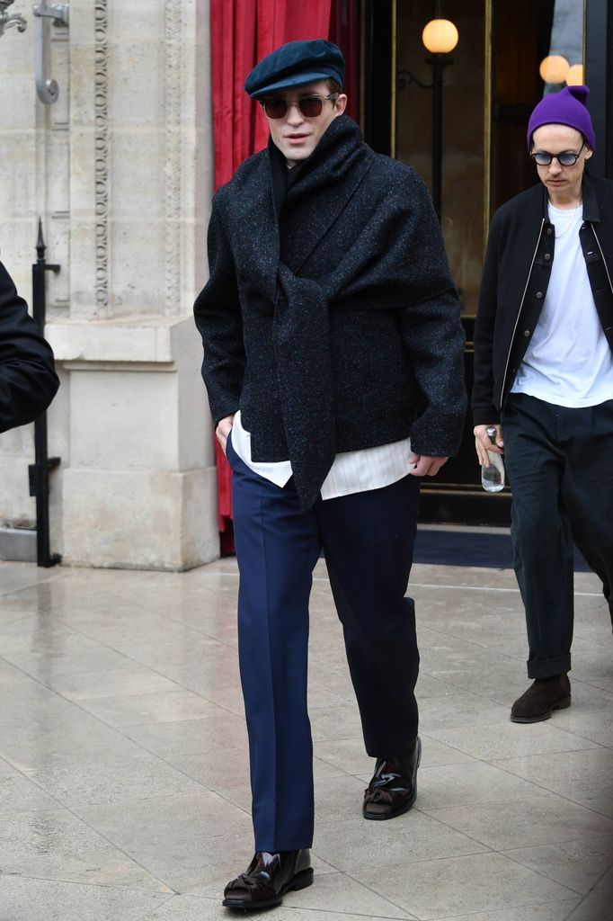 Robert Pattinson wore a textured navy coat with an oversized scarf, sharp blue trousers, a beret, and sunglasses, embodying intellectual chic with a Parisian twist.