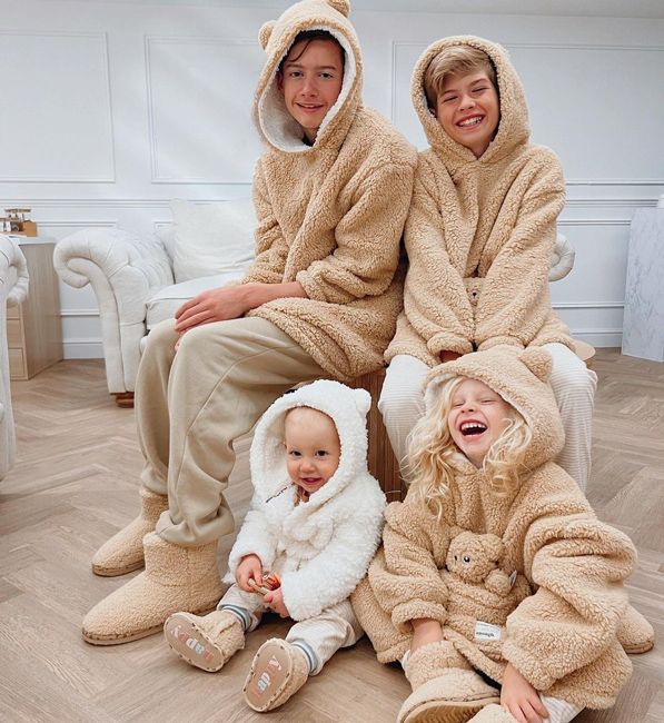 two teenage boys sit on a bench with a toddler and baby sitting on the floor in front of them all smiling and wearing coordinating fluffy beige and cream onesies in a neautral coloured room