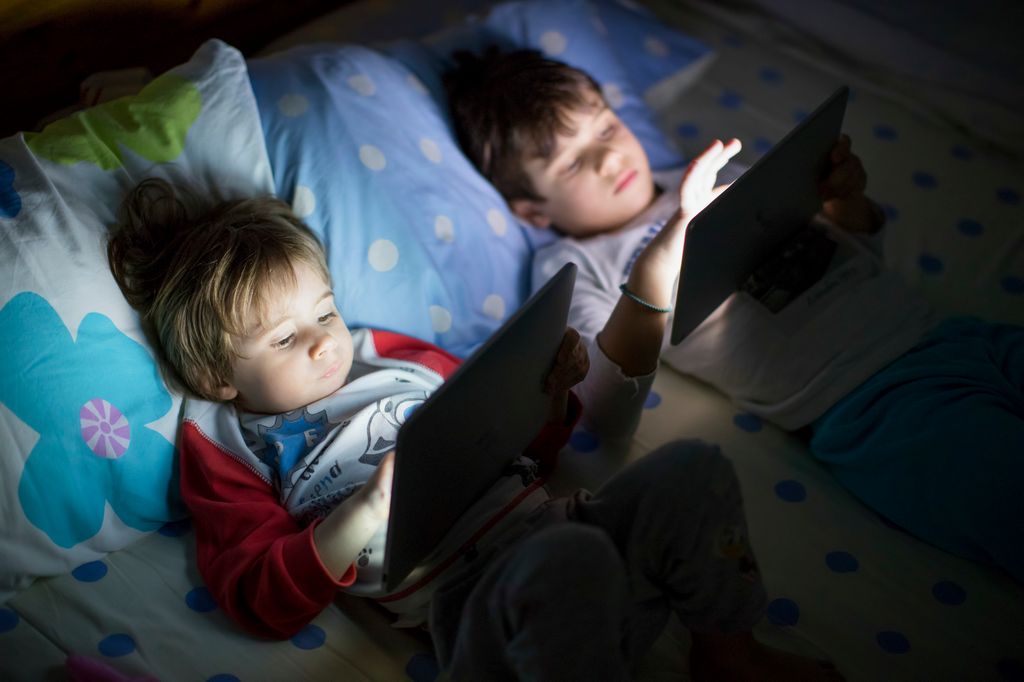 In a cozy bedroom, two children lie on a bed, focused on tablets in dim light. One child is intently reading while the other watches a screen, embodying nighttime routines of modern families.
