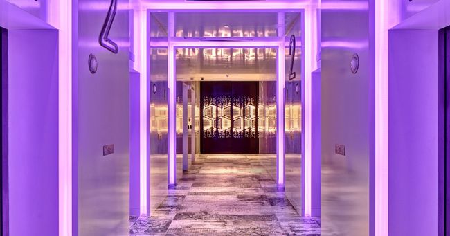 a marble floored corridor lit with lilac neon pannels with numbered bedroom doors on either side