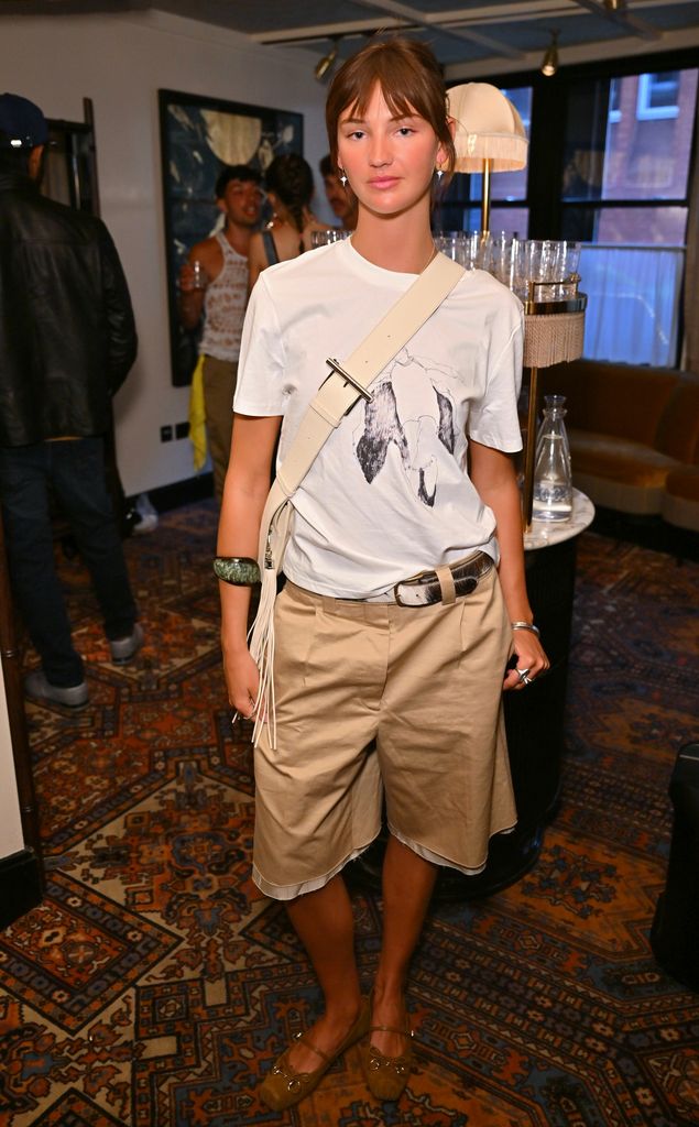 Mia Regan attends the Choose Love T-shirt launch in London wearing khaki Bermuda shorts, a graphic white tee, a cow-print belt and ballet flats, styled with a white crossbody and sculptural accessories.