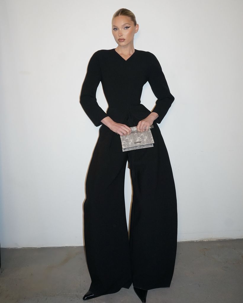 In the image, Elsa stands poised against a plain white backdrop, allowing her sleek black outfit to command full attention. She wears a sharply tailored, V-neck black jacket, cinched subtly at the waist, accentuating her silhouette without resorting to overt drama. The jacket gives way to voluminous, wide-leg trousers that billow and skim the floor, creating a statuesque effect. Her hair is parted in the center and slicked back into a polished, low bun, highlighting her flawless, contoured features and winged eyeliner. In one hand, she clutches a textured, snakeskin-print clutch, adding a dash of tactile intrigue to her otherwise monochrome palette. The look is finished with pointed black heels just peeking beneath the generous hem of her trousers.