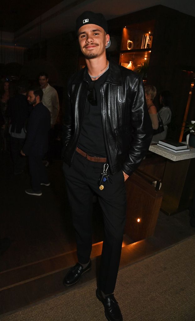 Romeo Beckham  in a black leather jacket, matching pants, and a black baseball cap stands confidently with a smile. His casual-chic look includes a chain necklace and key accessories.