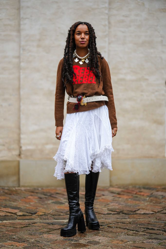 A guest wears a white large pearls with silver pendants necklace, a dark brown with embroidered red pattern pullover, a white latte matte leather belt, a white ruffled knees skirt, black shiny leather block heels / platform knees boots / tight high waders boots , outside Marimekko, during the Copenhagen Fashion Week Spring/Summer 2024 