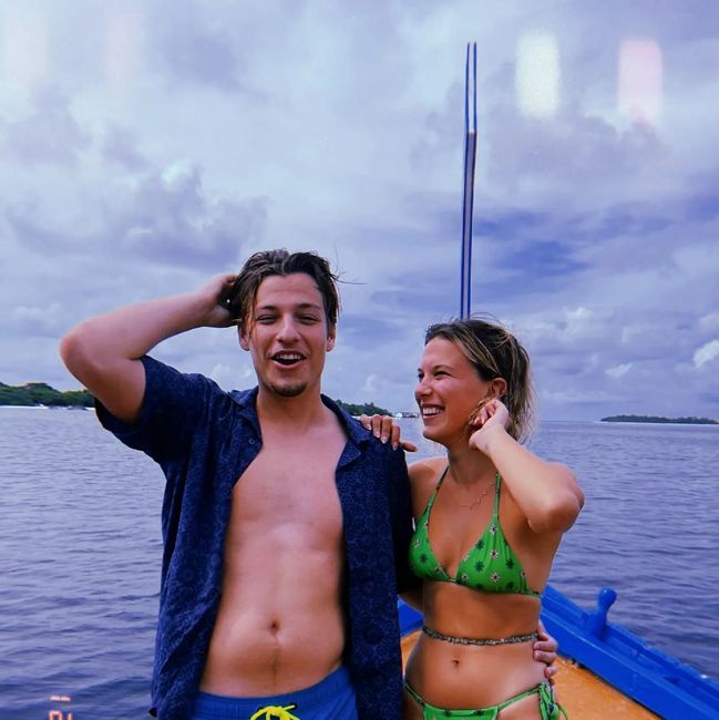 a man stands on a boat at sea with an open shirt as a woman stands beside him wearing a green bikini and smiles at him as he brushes his hair back with one hand