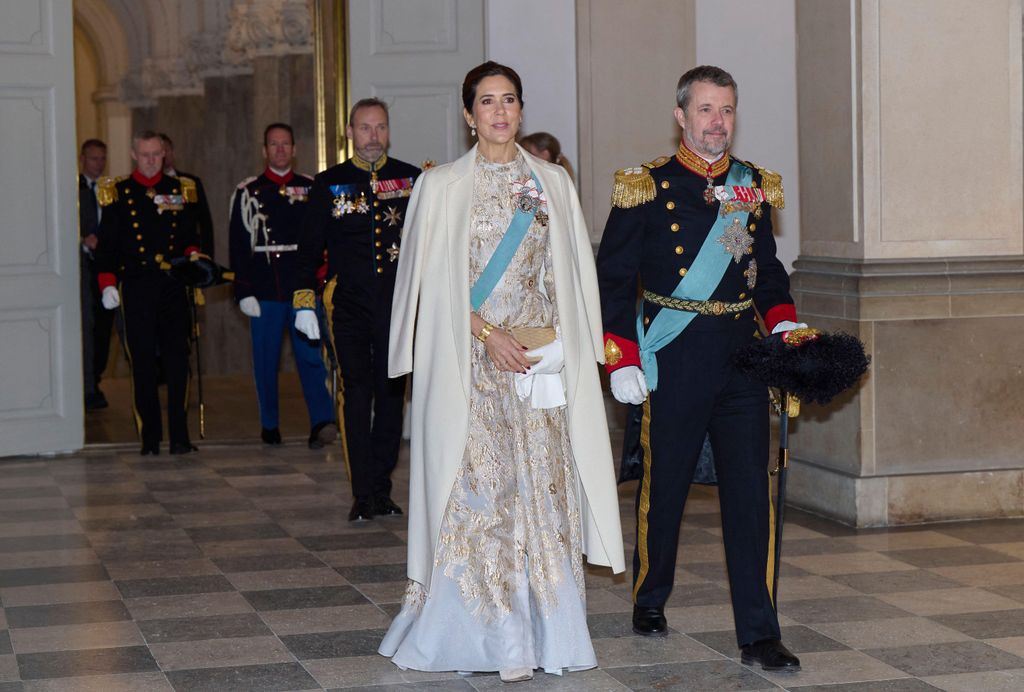 King Frederik in uniform and Queen Mary in white dress walking through palace hall