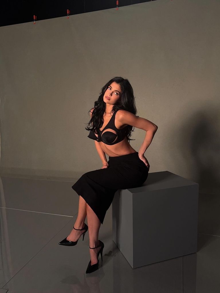 In a moody studio setting, Kylie Jenner sits confidently on the edge of a matte grey block against a neutral background. She wears a sculptural black bra top that dramatically contours and lifts, featuring a striking cutout design reminiscent of Madonna’s iconic cone bra era. Her voluminous dark hair cascades in soft waves, framing a glamorously contoured face with a nude-pink pout. A high-waisted black skirt hugs her curves and flows just below the knee. Her manicured nails are long, sharp, and white — a modern femme fatale finishing touch. The lighting creates subtle shadows, lending a cinematic quality to the image, as if Kylie has just stepped off a 90s runway or into a modern-day fashion noir.