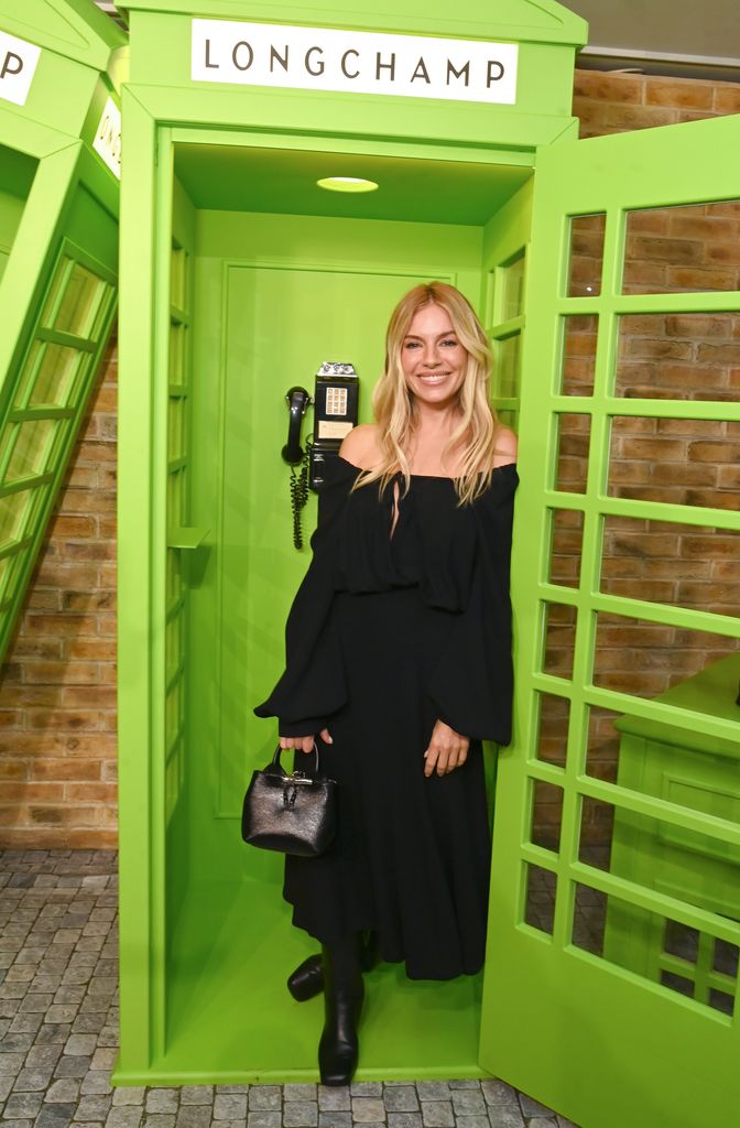 Sienna Miller wearing an off-the-shoulder black dress while standing in a neon green photobooth