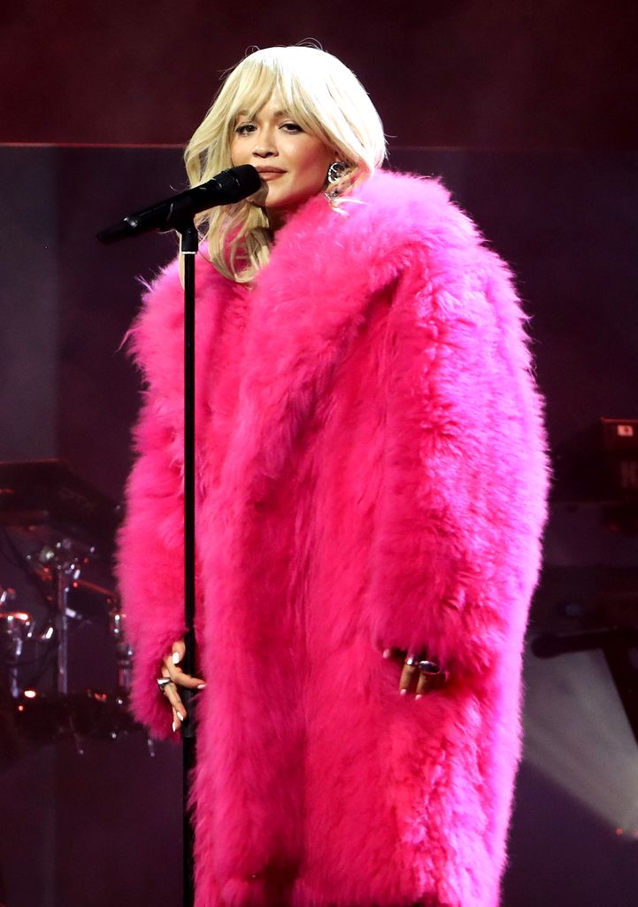  Rita Ora performs onstage during the Music Industry Trust Awards 2025 wearing a huge pink fluffy coat. She looks to the camera and brushes the microphone stand with one hand. 