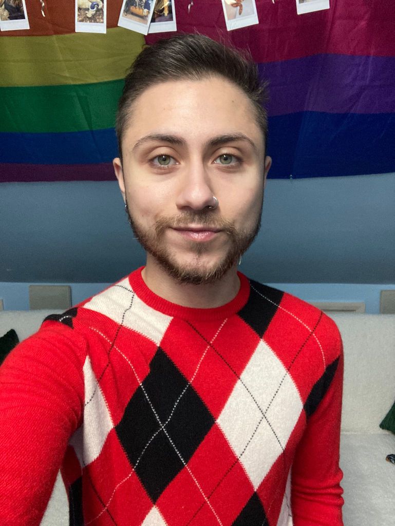 A man with facial hair in a black and red jumper with a pride flag and bisexual pride flag hanging on the wall
