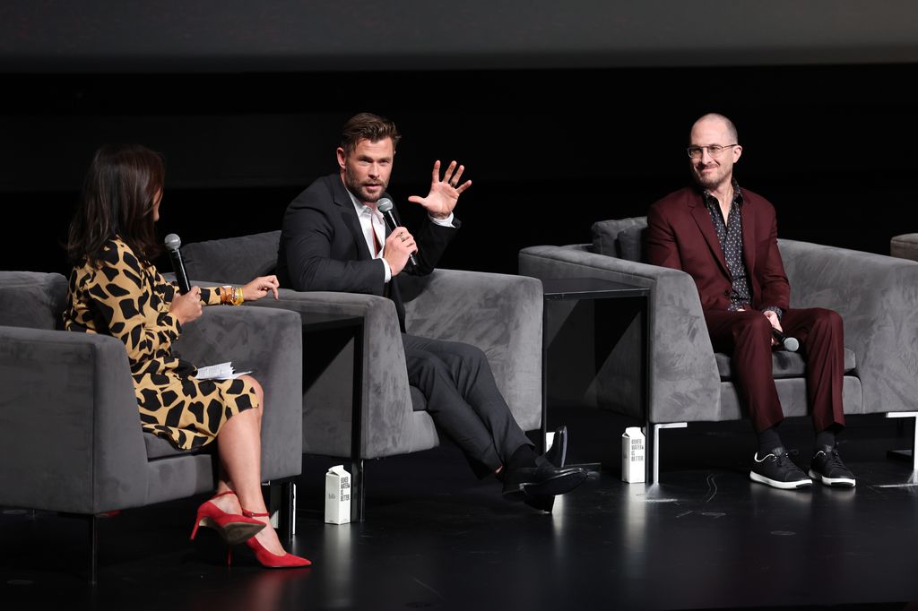 Chris Hemsworth sat on a stage being interviewed by another person, who has her back to the camera