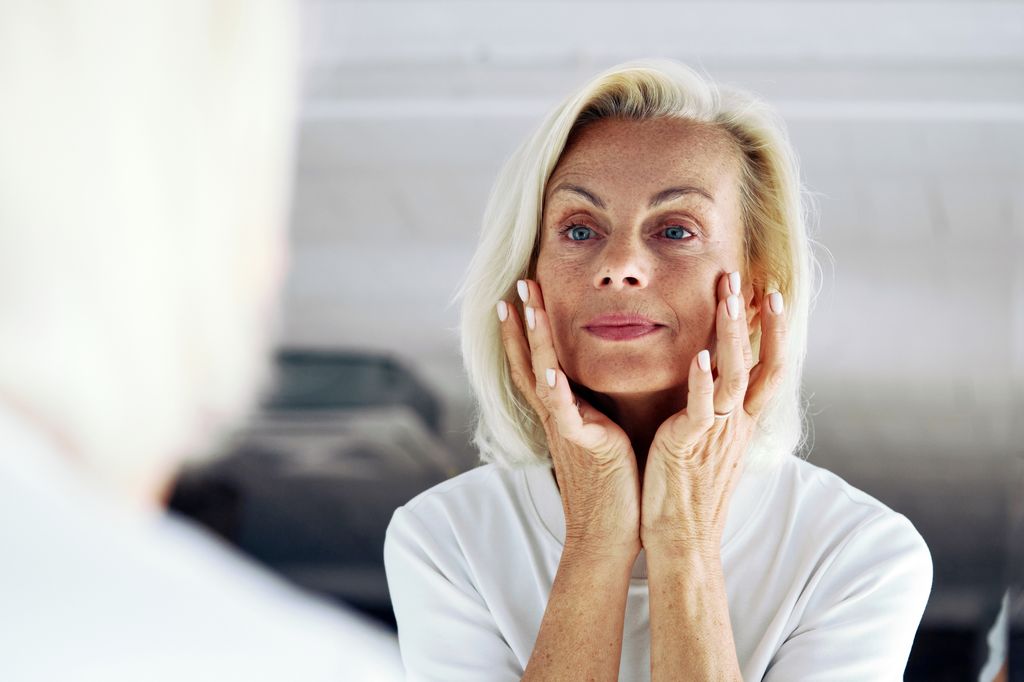 Close up of older 65s woman stand in front of mirror, gently touch her face with hands, examining skin, assessing results of skincare routine. 