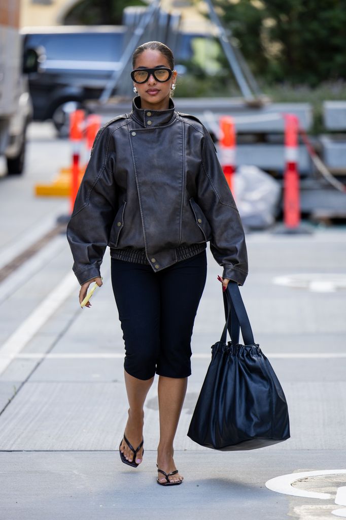 A guest wears black bag, brown leather jacket, capri pants, sunglasses, flip flops outside Herskind during Copenhagen Fashion Week day three on August 06, 2025 in Copenhagen, Denmark. 